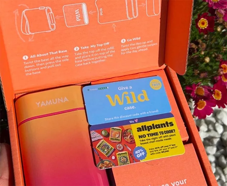 Open orange box contains a pink and orange insulated bottle with the brand name "Yamuna." Accompanying it are promotional cards offering discounts for "Wild" and "allplants" products. Flowers are in the background.