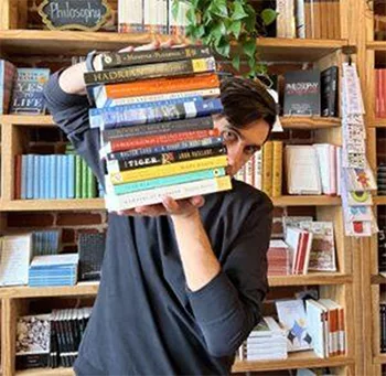 A person in a bookstore holds a tall stack of books with both hands, obscuring part of their face. Shelves filled with various books are visible in the background.