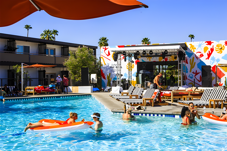 People relaxing and swimming in a clear blue pool surrounded by lounge chairs and vibrant murals on a sunny day.