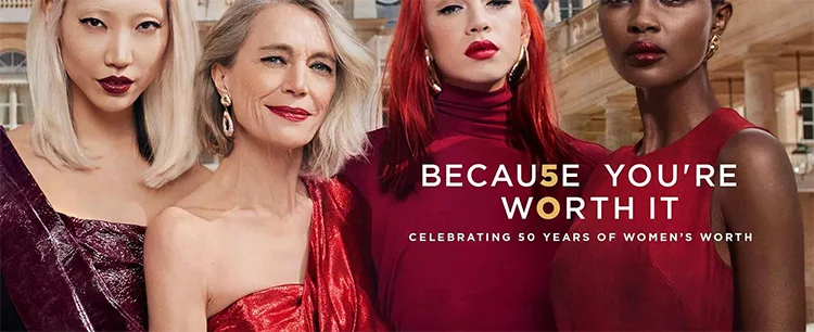 Four women, each with distinctive hairstyles and makeup, wearing elegant red and pink outfits, stand together against an ornate background. Text reads: "Because You're Worth It. Celebrating 50 Years of Women's Worth.