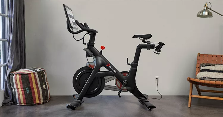 A stationary exercise bike with a screen is set up in a minimalist room with a striped ottoman and a wooden chair.