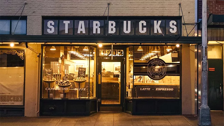 The front of a Starbucks coffee shop at night with lights on, showing large glass windows and a door displaying the menu items "Cappuccino," "Coffee," "Latte," and "Espresso.