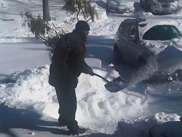 A person is shoveling snow next to a parked car on a snowy street.