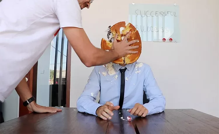 A person in a white shirt smashes a cake into the face of another person in a blue shirt and tie sitting at a table with a "Successful Success" sign on the wall in the background.