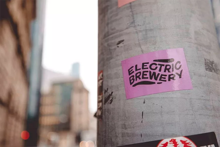 A purple sticker with the text "Electric Brewery" is on a gray utility pole in an urban setting.