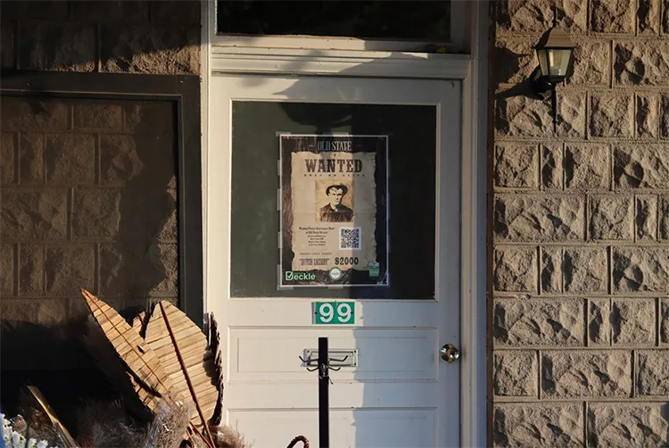 A wanted poster featuring a man's photo is taped to a white door with a glass window. The door is flanked by a wall lamp and has a green house number "99" displayed.