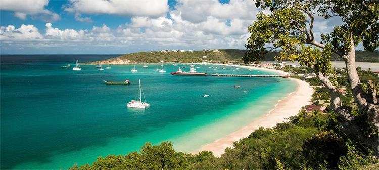Aerial view of a bay with turquoise water and sandy beach, dotted with anchored boats, surrounded by lush greenery under a partly cloudy sky.